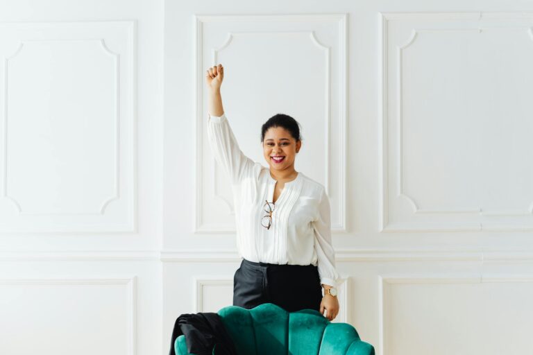 Confident woman in white blouse celebrating success with raised arm indoors.